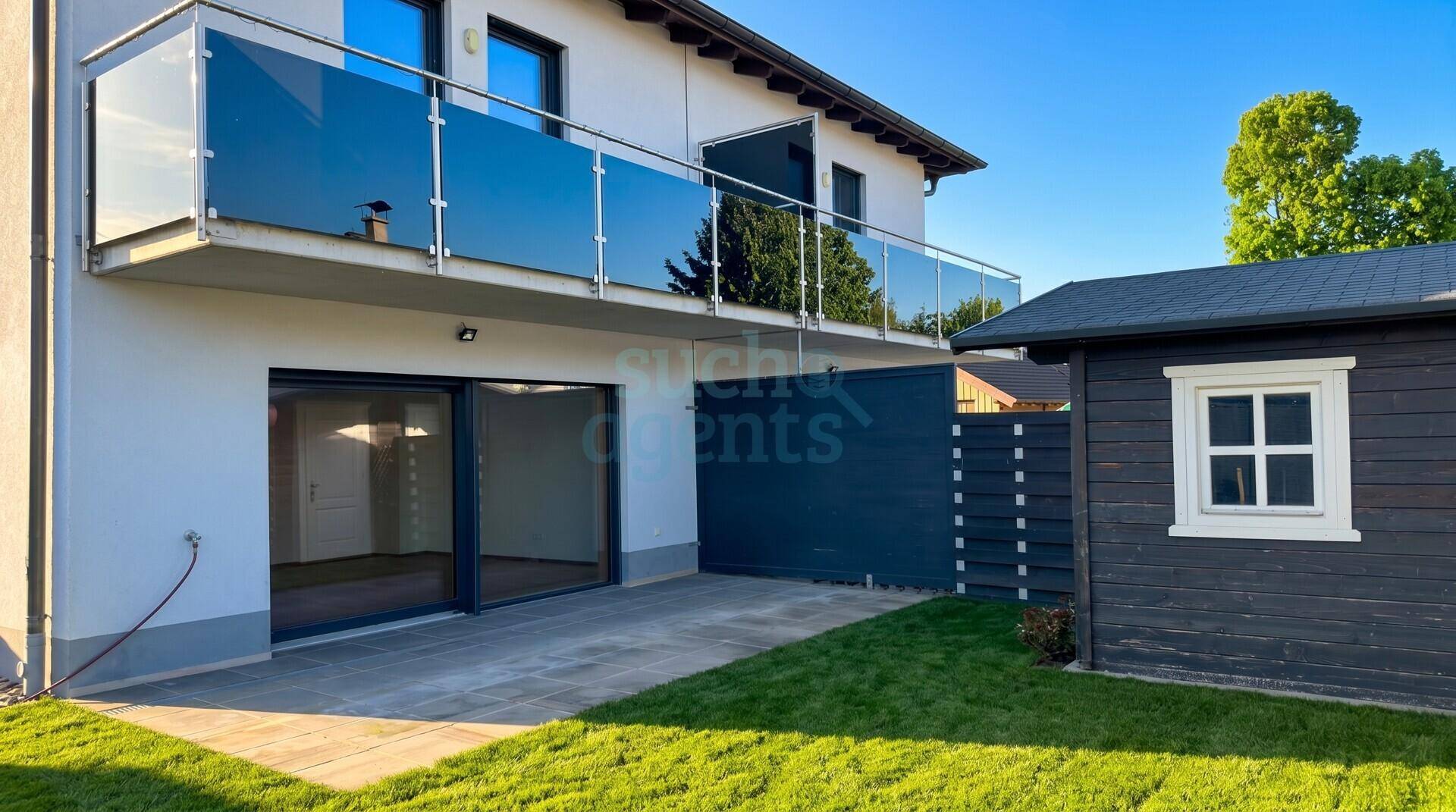 Außenansicht des Hauses mit Balkon, gepflasterter Terrasse und grünem Rasenbereich.