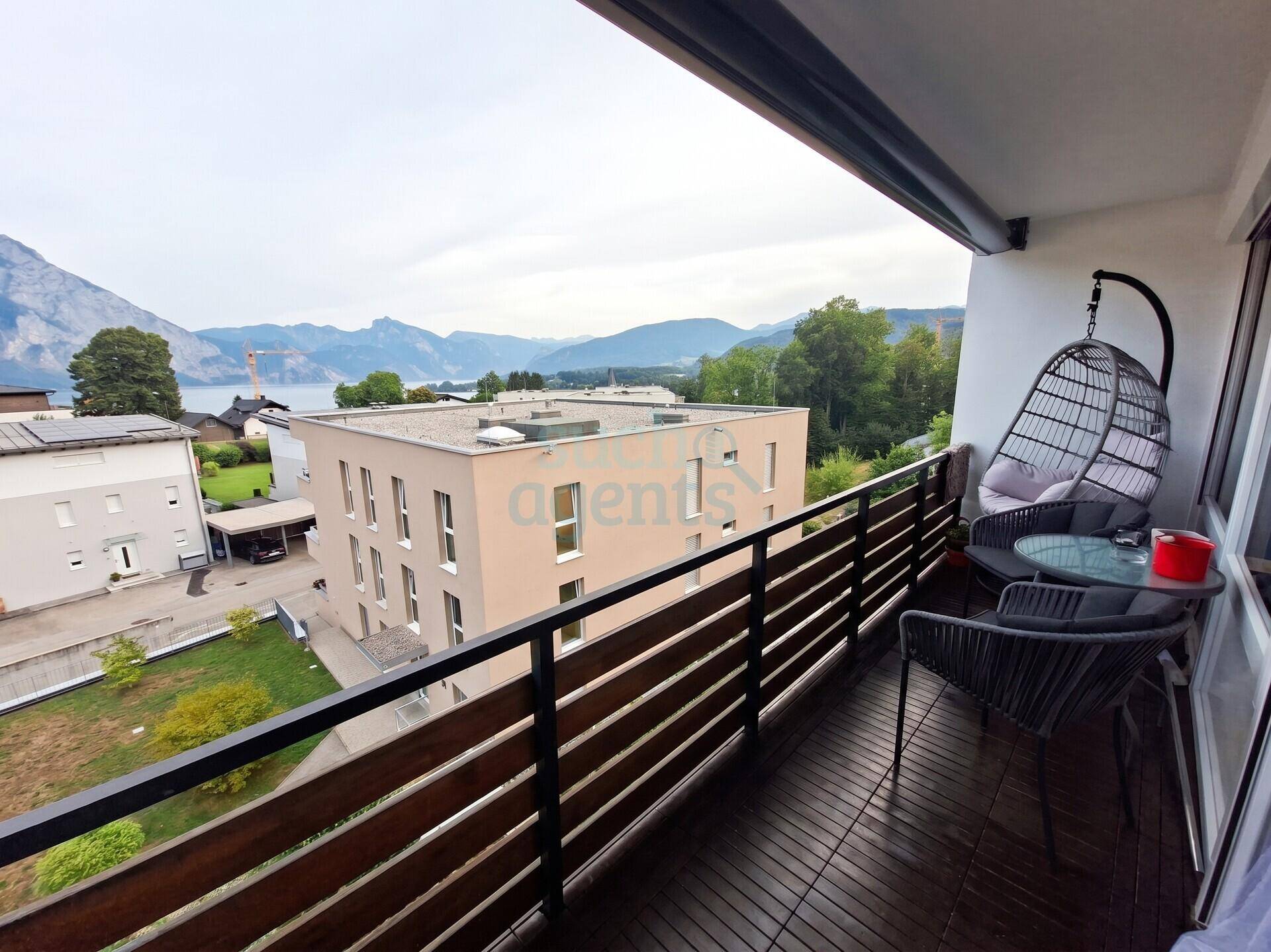 Geräumiger Balkon mit Holzboden, modernen Sitzgelegenheiten und malerischem Blick auf den See und die Berge.