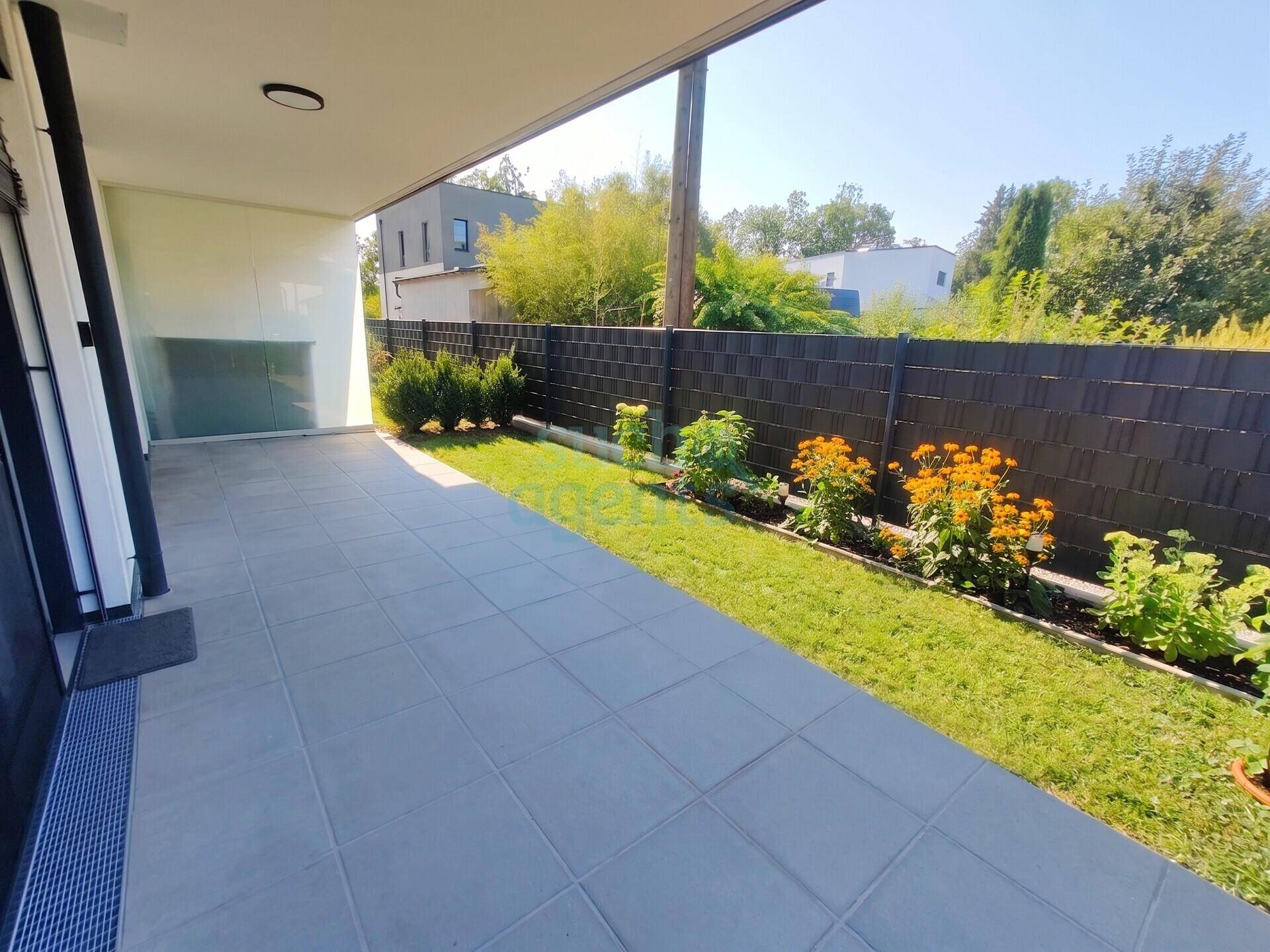 Geflieste Terrasse mit kleinem Rasenstreifen und Blick auf den Gartenbereich.