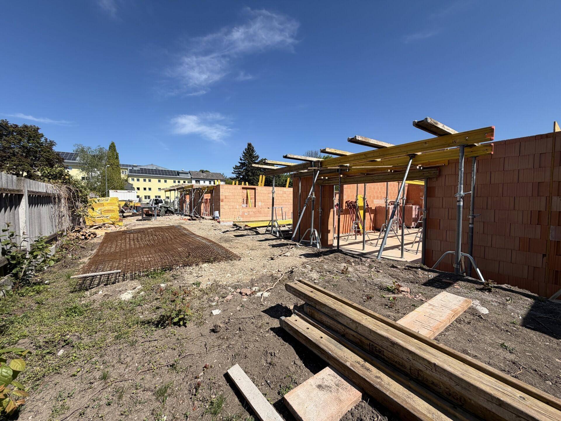 Baustelle eines Neubaus mit bereits errichteten Ziegelwänden und sichtbaren Solaranlagen im Hintergrund.