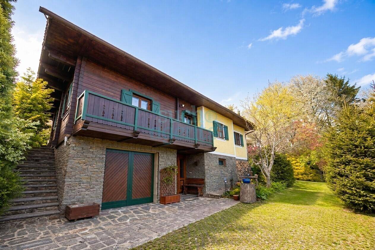 Traditionelles Holzhaus mit Steinfassade, Balkon und großem Gartenbereich unter blauem Himmel.