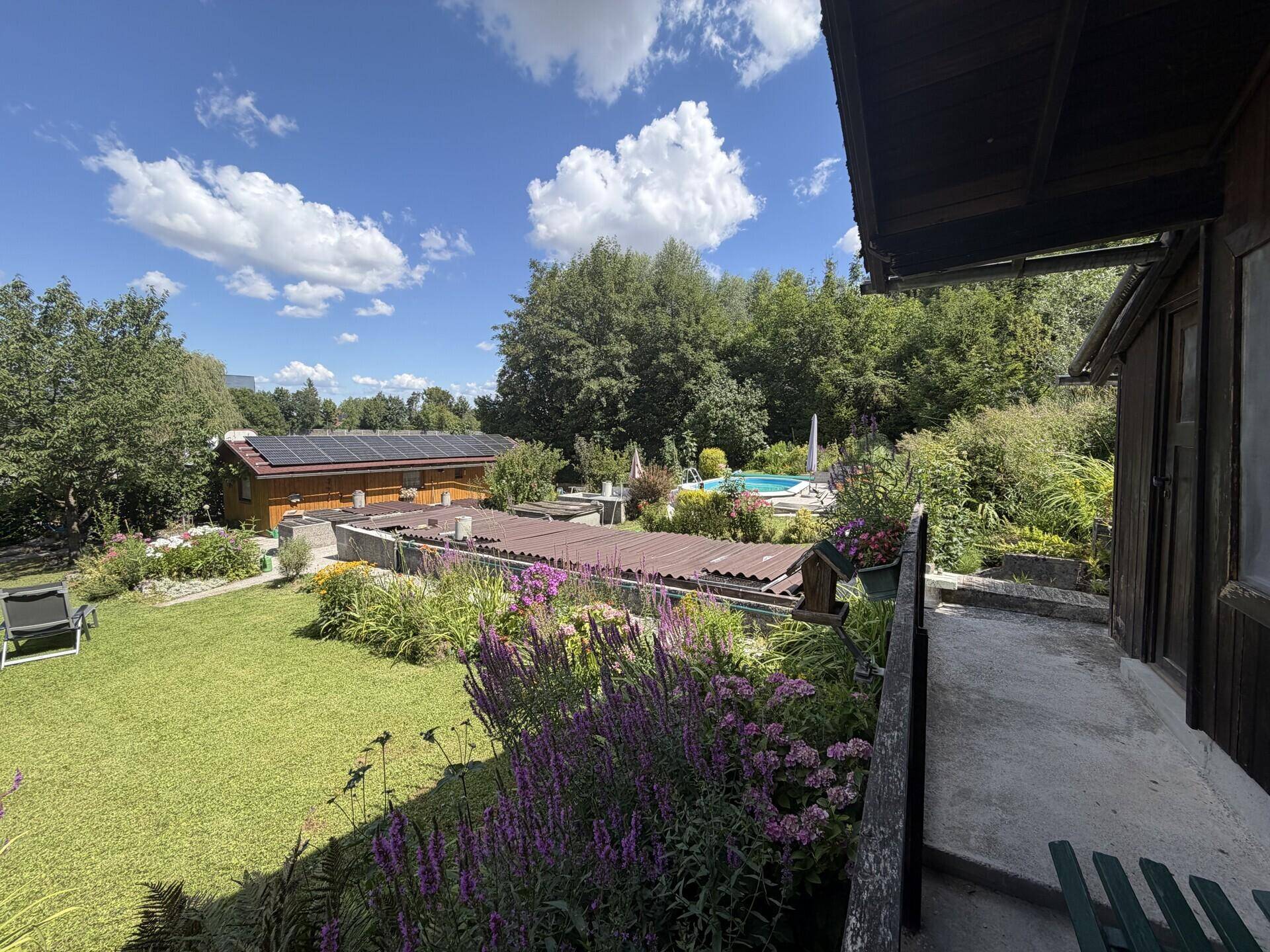 Weitläufiger Garten mit Rasenfläche, Blumenbeeten und einem Pool im Hintergrund unter blauem Himmel.