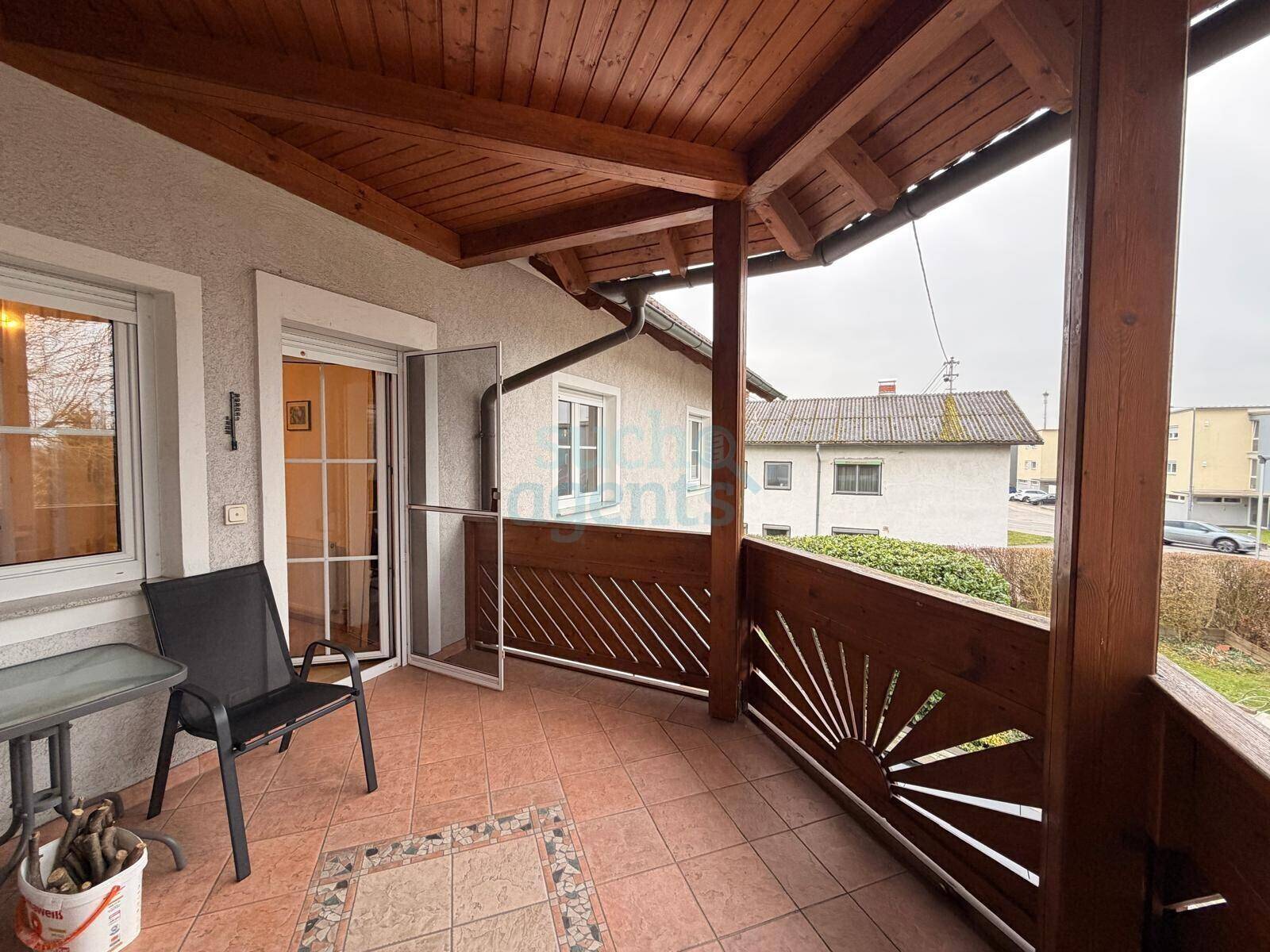 Überdachter Balkon mit Fliesenboden, Holzgeländer und Blick auf die Umgebung.