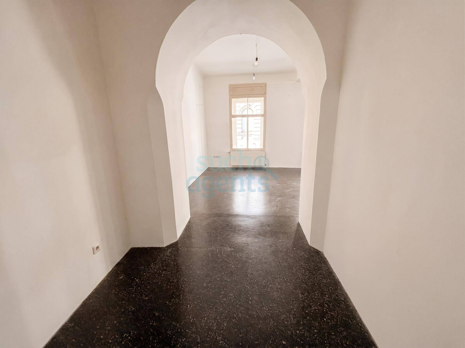 Blick durch einen gewölbten Flur in einen weiteren Raum mit dunklem Terrazzoboden und einem Fenster.