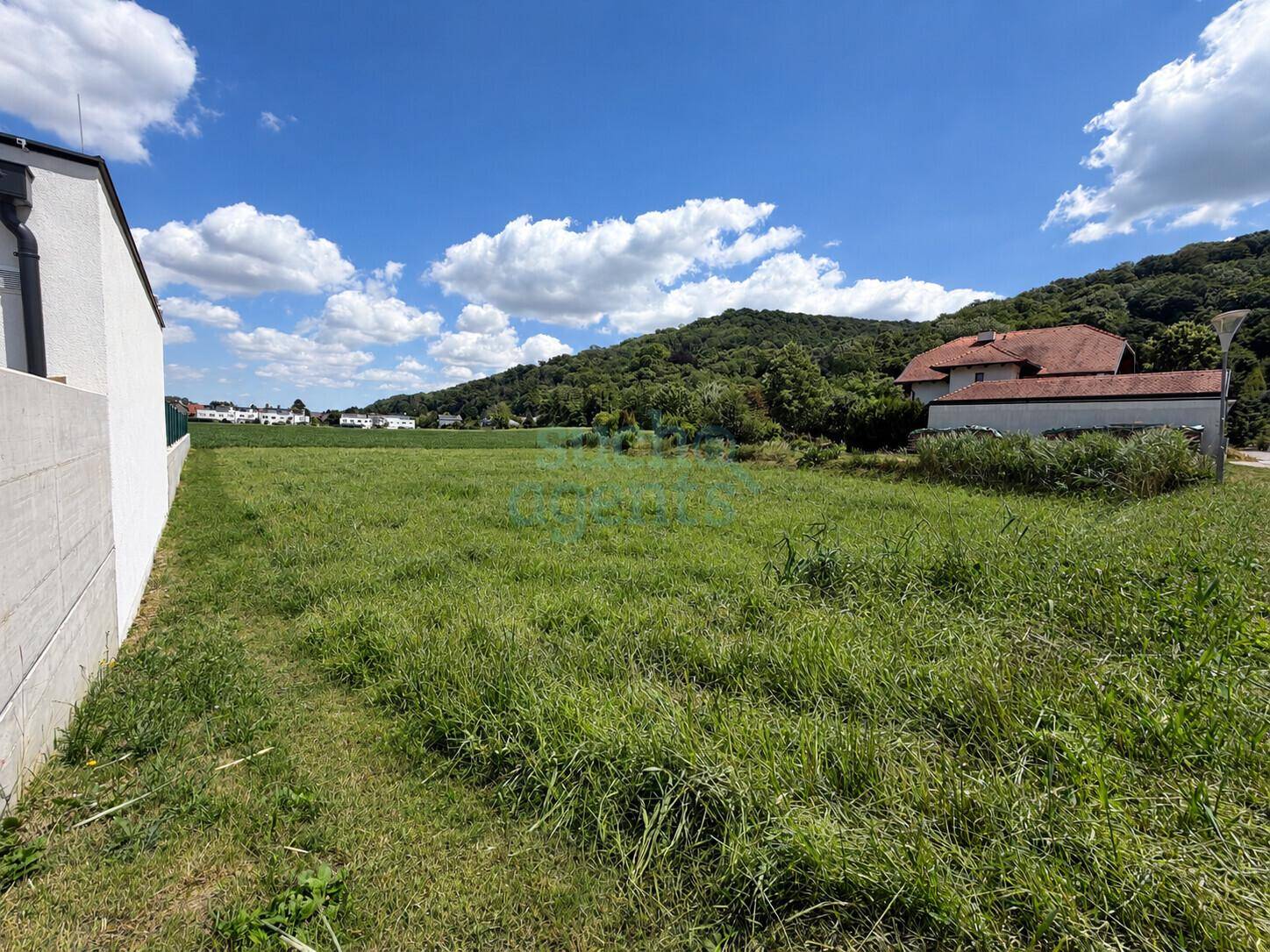 Grünes Grundstück mit einem modernen Gebäude im Vordergrund und bewaldeten Hügeln im Hintergrund.
