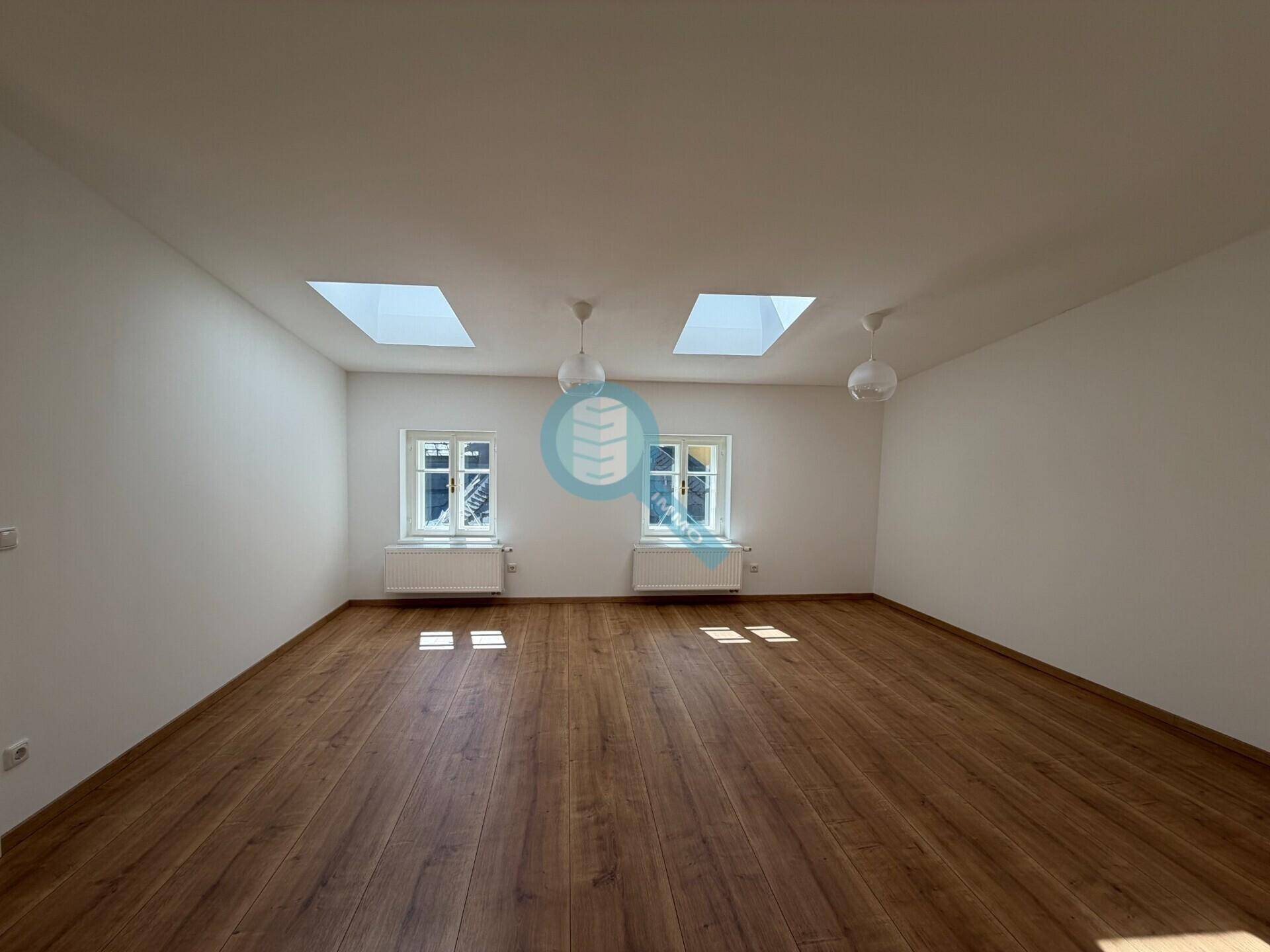 Geräumiger, leerer Raum mit Holzboden, weißen Wänden und zwei großen Dachfenstern.