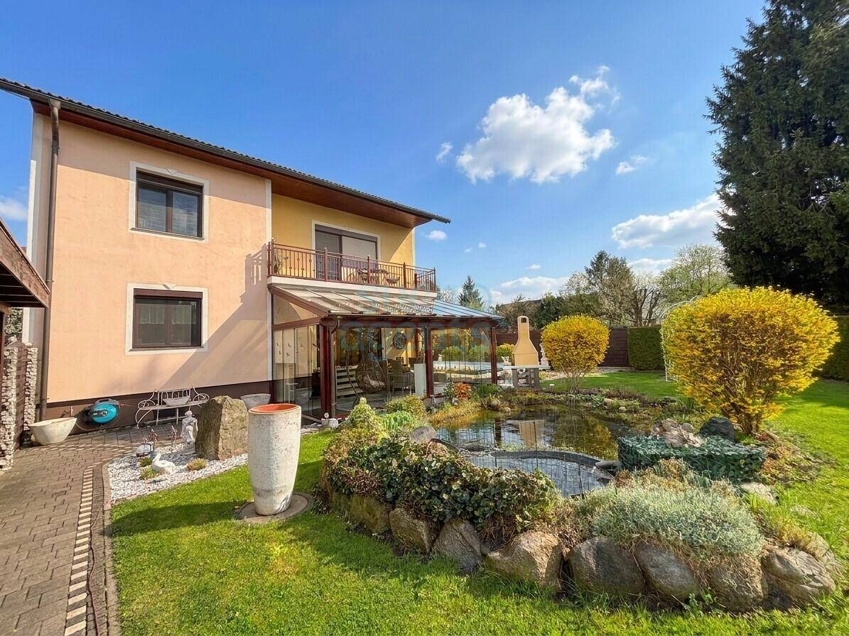 Gepflegter Garten mit Teich, Sonnenterrasse und Balkon am Wohnhaus unter blauem Himmel.