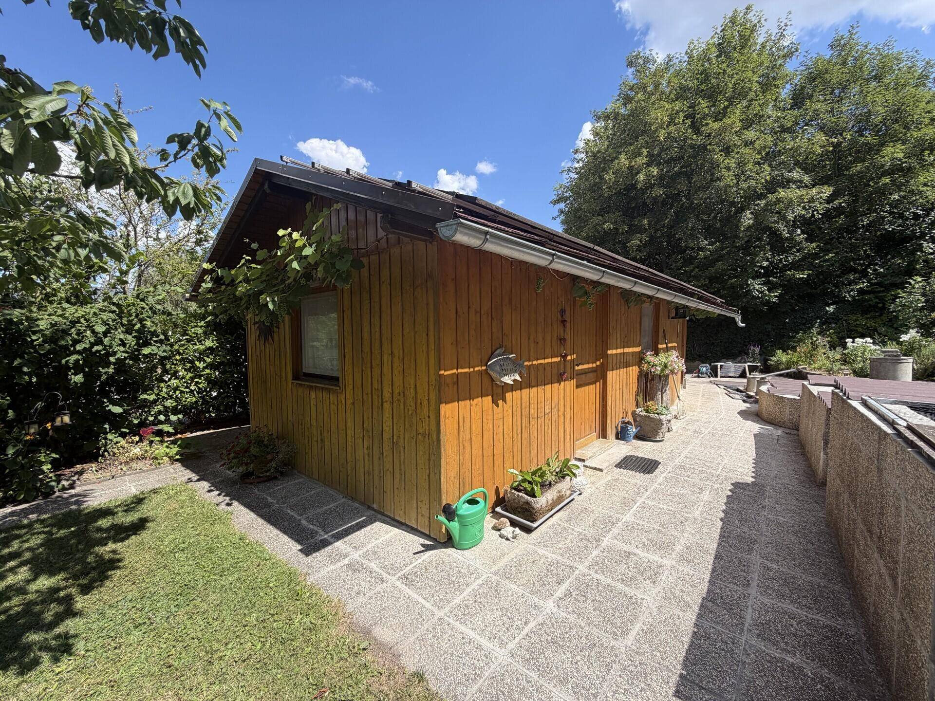 Kleines Holzhaus im Garten mit gepflastertem Weg und umgebender Grünfläche unter blauem Himmel.