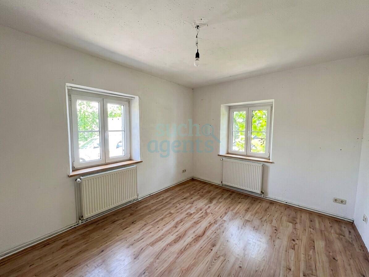 Helles Zimmer mit Holzboden, weißen Wänden und zwei Fenstern.