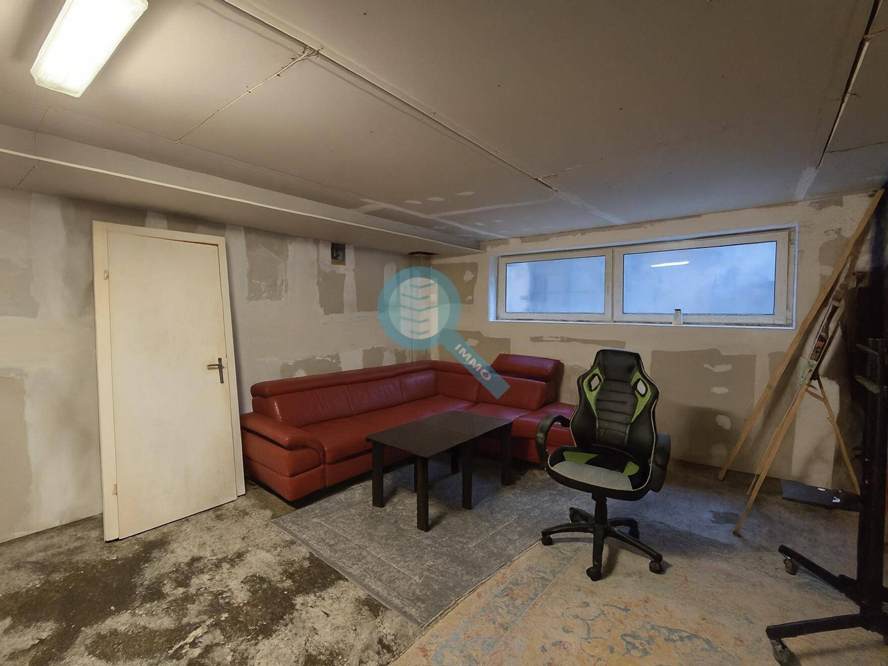 Wohnraum mit rotem Ecksofa, Couchtisch und Gaming-Stuhl, die Wände sind noch im Rohbauzustand.