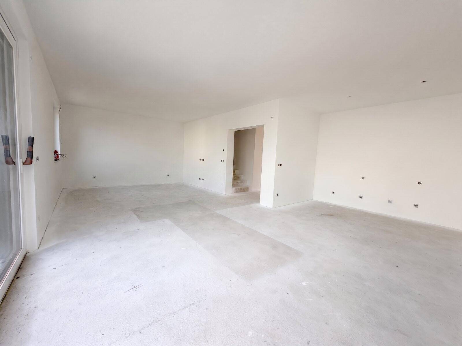 Geräumiger, unmöblierter Innenraum im Rohbau mit großen Fenstern und Betonboden.