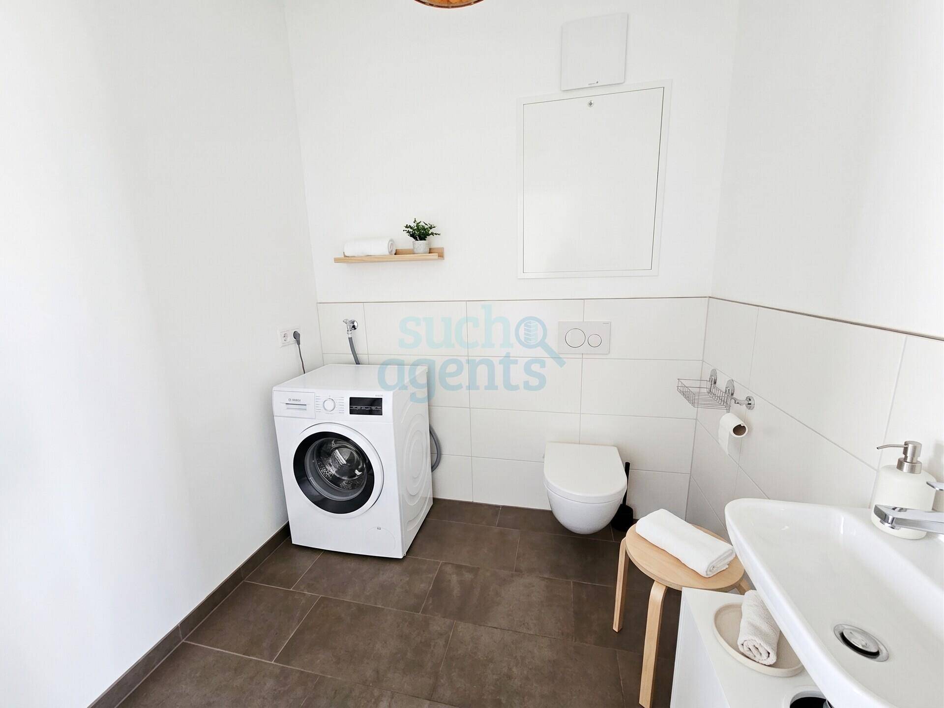 Funktionales WC mit Waschbecken, Toilette und integrierter Waschmaschine, hell gefliest.