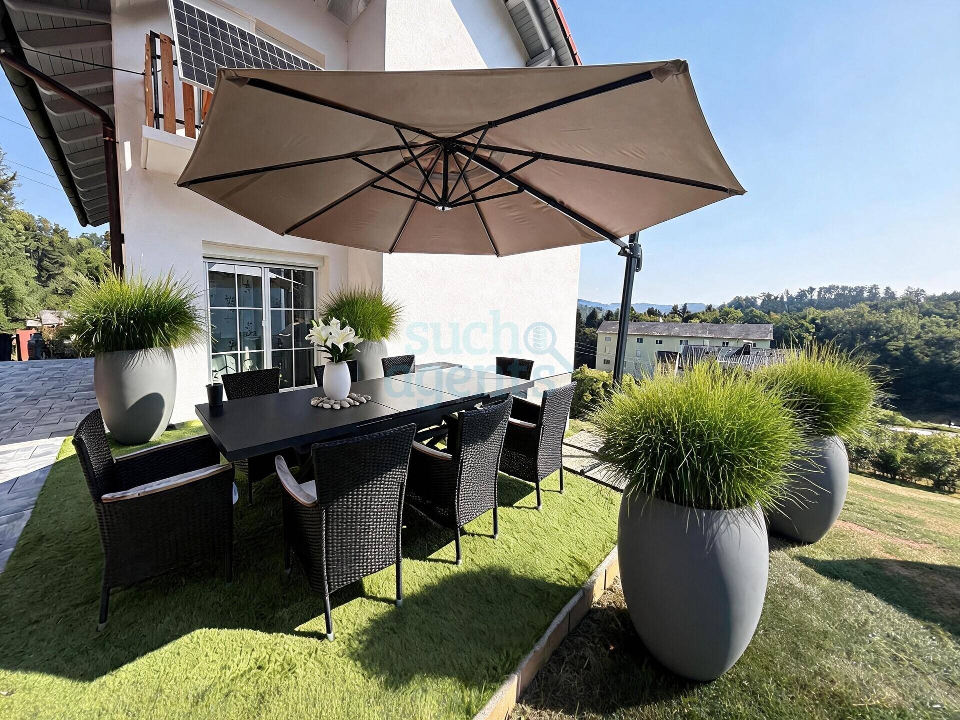 Sonnige Terrasse mit großem Esstisch, Stühlen, Sonnenschirm und Blick auf die grüne Landschaft.