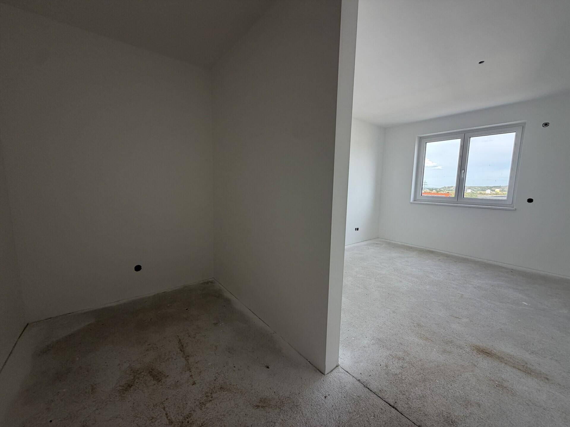 Unmöblierter Raum im Rohbau mit weiß gestrichenen Wänden, Betonboden und Fenster.