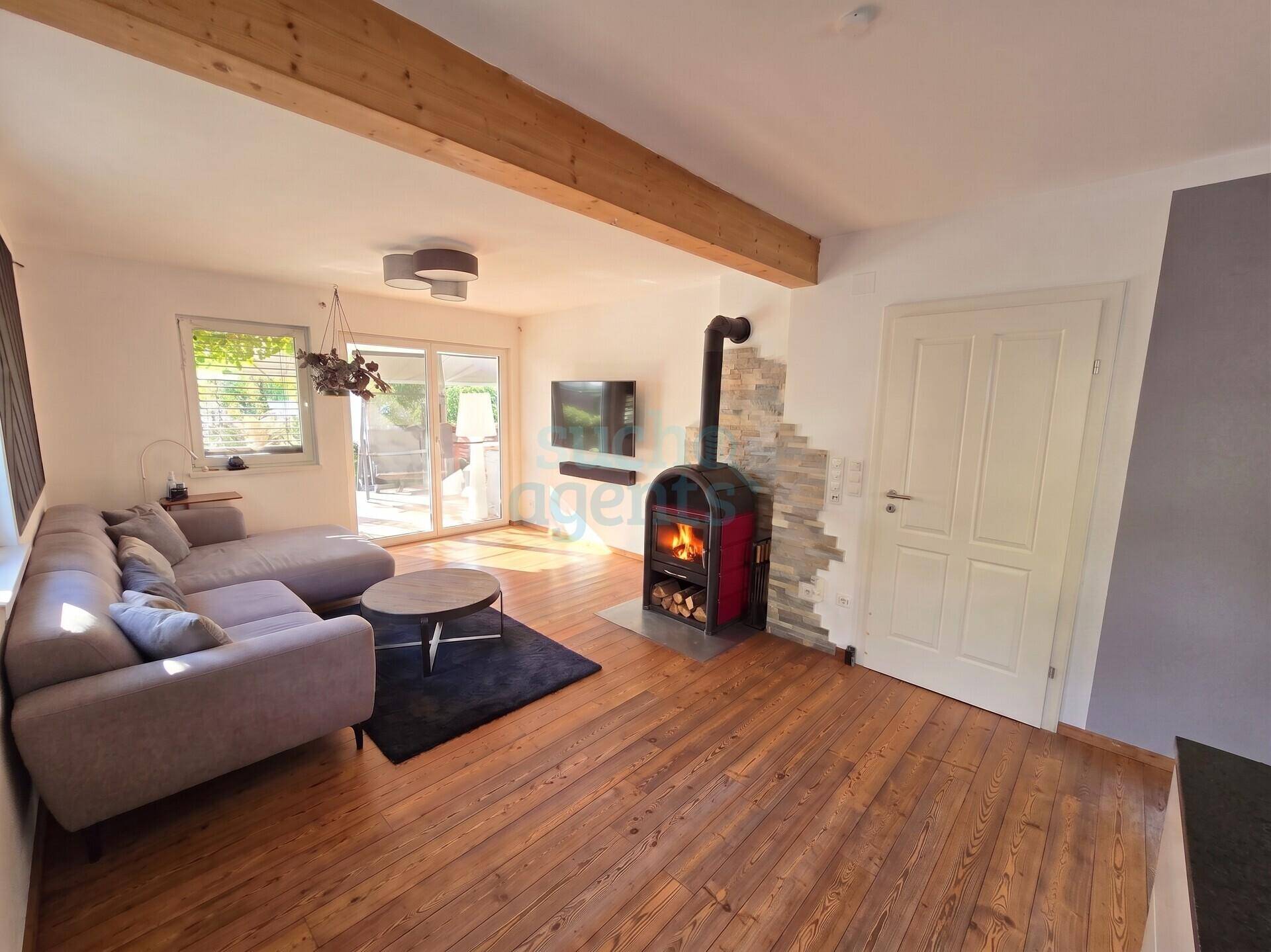 Gemütliches Wohnzimmer mit Holzboden, modernem Kaminofen und Zugang zur Terrasse, unter sichtbaren Holzbalken.