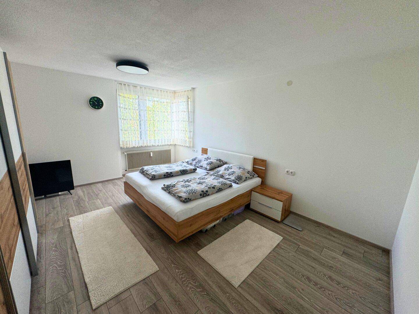 Schlichtes Schlafzimmer mit Holzbett, Nachttischen und einem großen Fenster für viel Tageslicht.