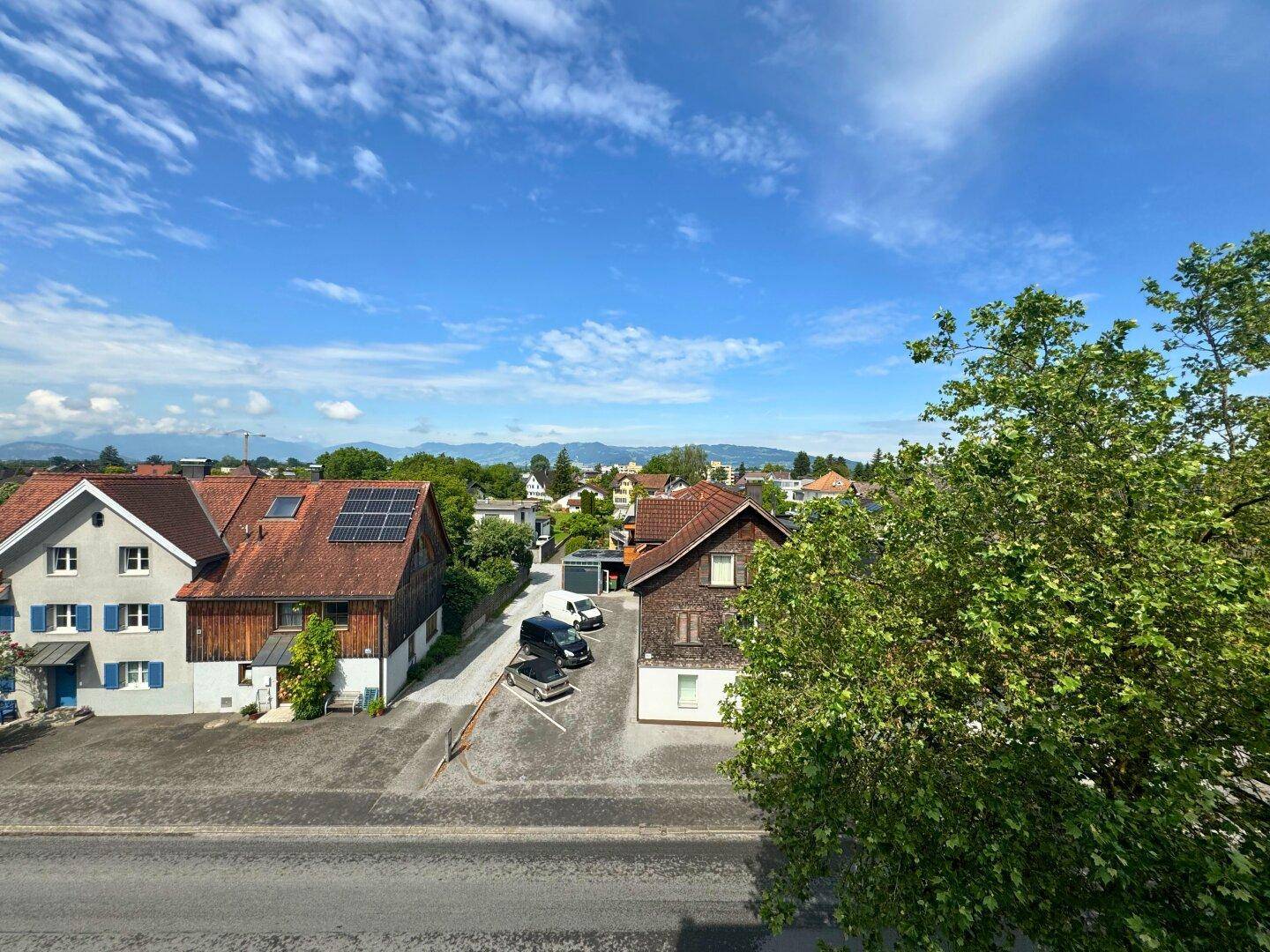 Weitblick über eine Wohnsiedlung mit Häusern, Bäumen und Bergen im Hintergrund unter blauem Himmel.