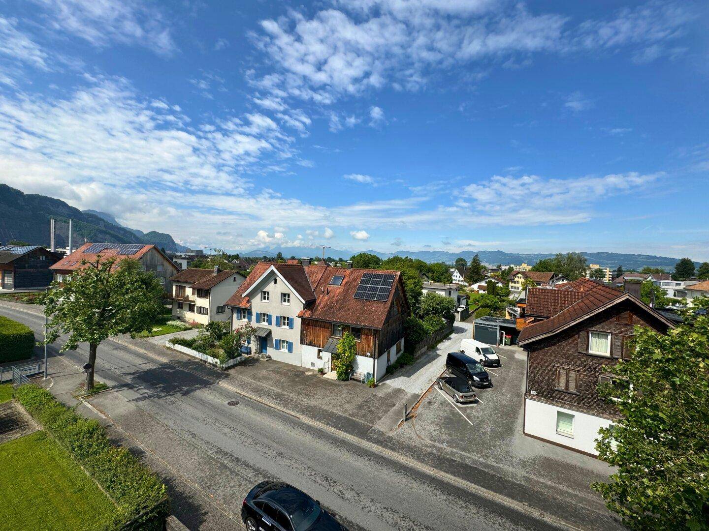 Panoramablick auf eine ländliche Wohngegend mit Häusern, grünen Bäumen und Bergen am Horizont.
