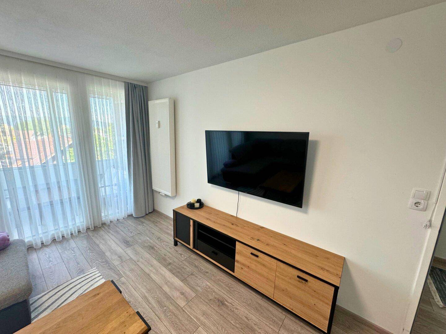 Wohnzimmer mit Holzboden, Wandfernseher und einem modernen Sideboard unter großen Fenstern.
