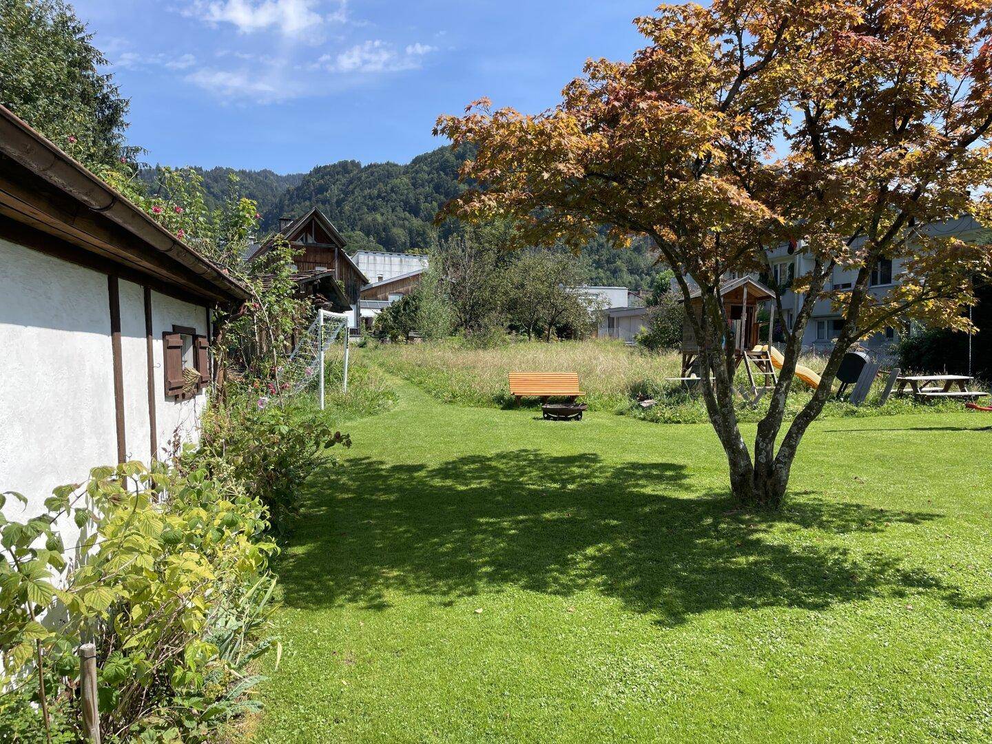 Gepflegter Gartenbereich mit Rasenfläche, einem Baum und einem Spielplatz im Hintergrund, umgeben von Bergen.