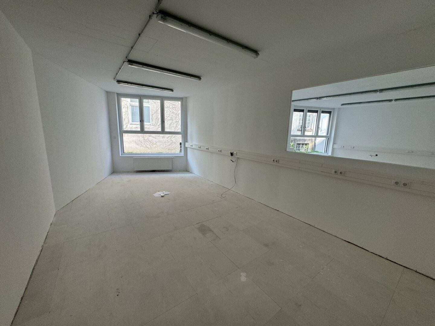 Leerer Raum mit einem großen Wandspiegel, Fenstern und einer Reihe von Steckdosen entlang der unteren Wand.