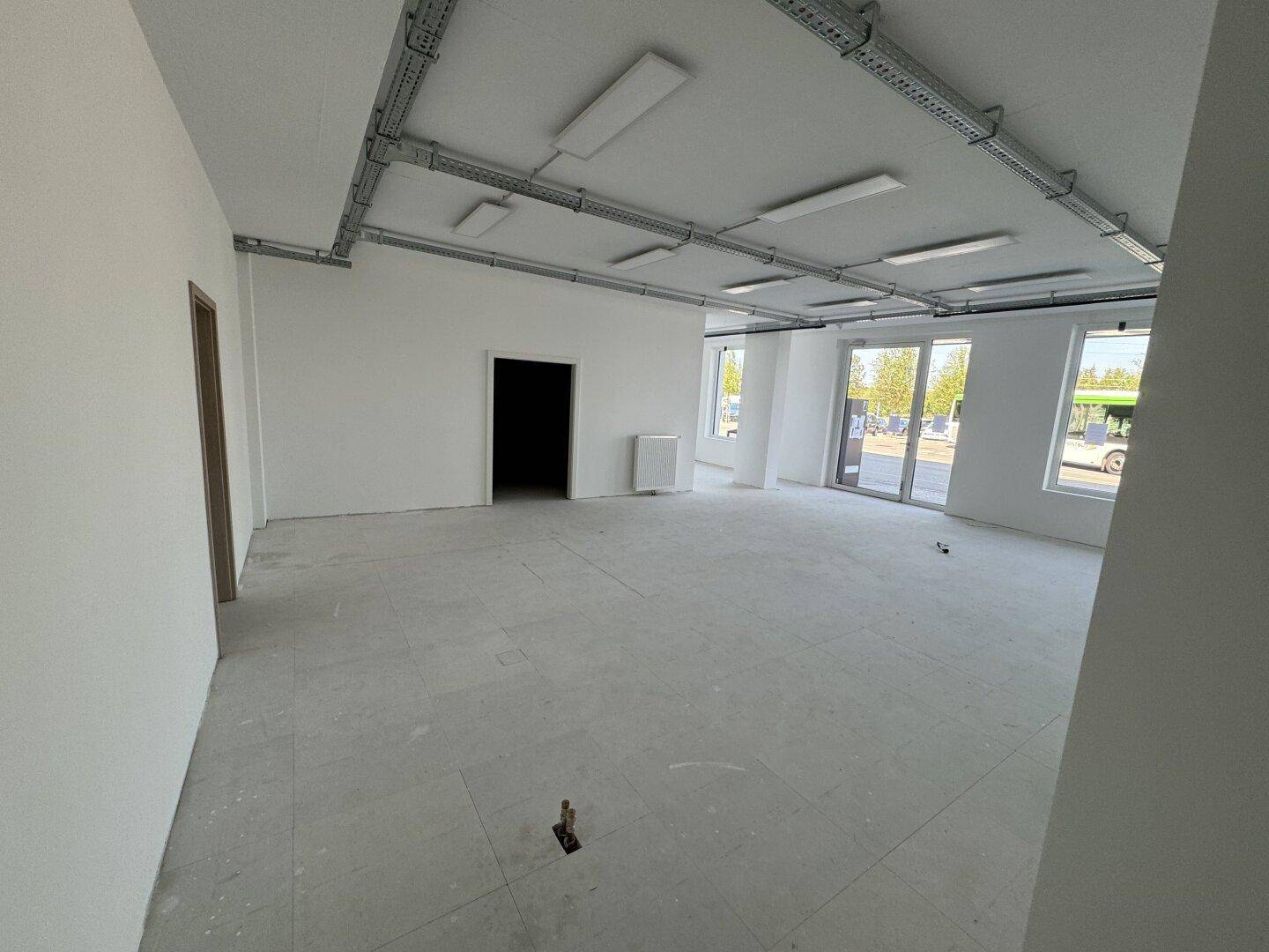 Großer, leerer Büroraum mit sichtbaren Deckeninstallationen, großen Fenstern und einem hellen Fliesenboden.