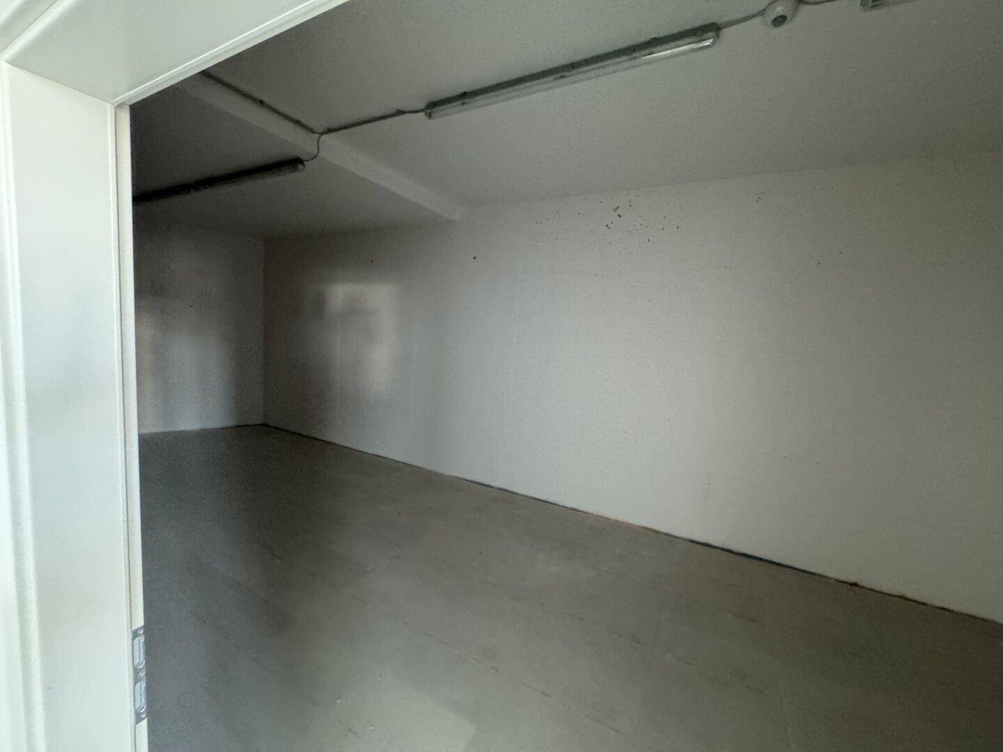 Langer, schmaler Lagerraum mit weißen Wänden und einem einfachen Boden, beleuchtet von Deckenleuchten.