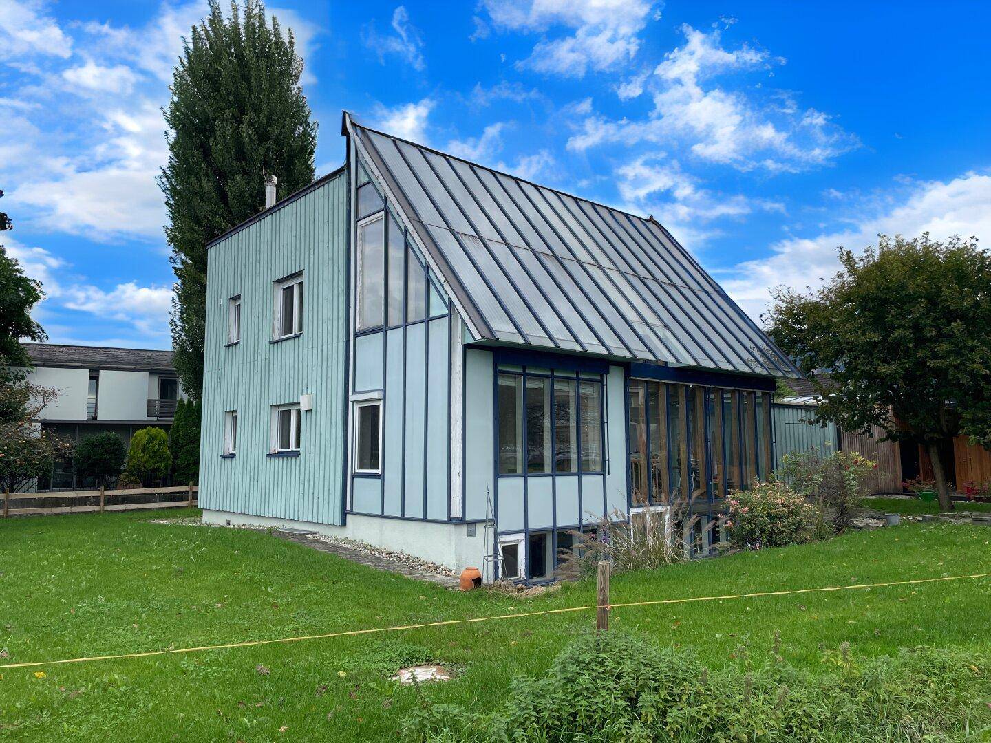 Modernes Haus mit hellblauer Holzfassade, großflächiger Verglasung und Satteldach in grüner Umgebung.
