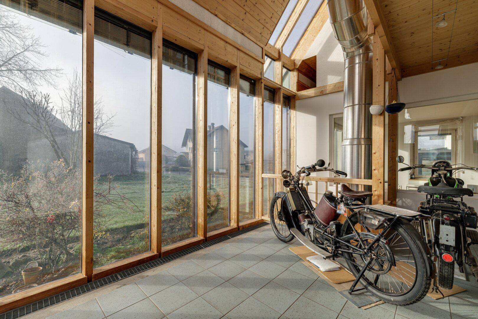 Heller Wintergarten mit großen Glasfronten, Holzbalken und Fliesenboden, ideal für Motorräder.