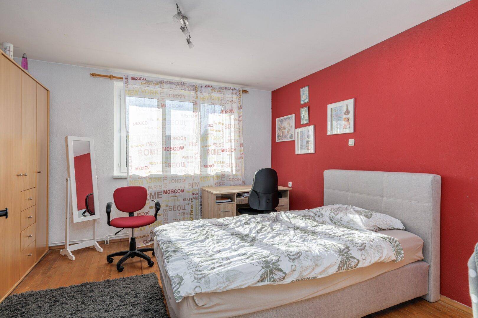 Schlafzimmer mit roter Akzentwand, Doppelbett und Schreibtisch am Fenster.