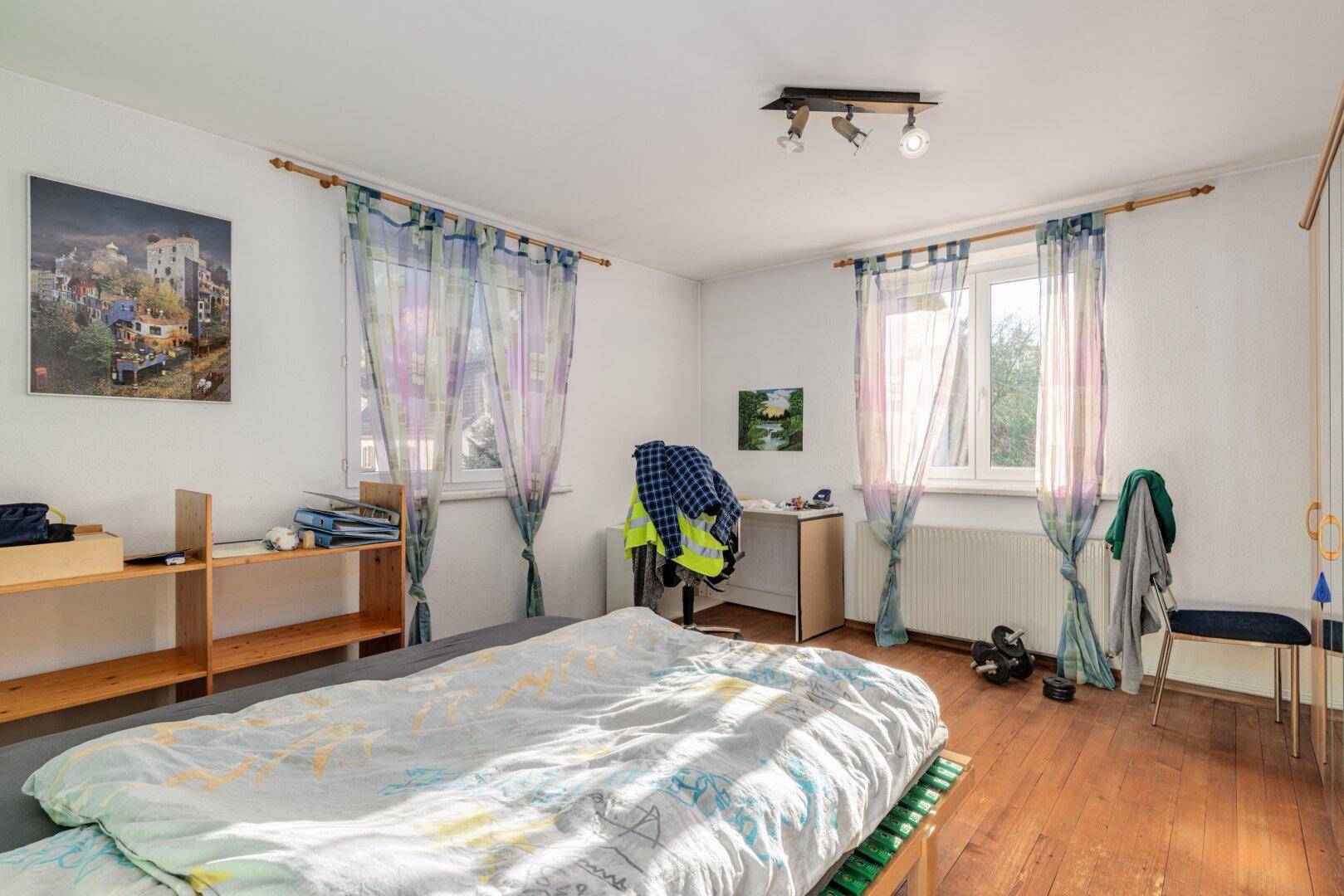 Schlafzimmer mit Holzbett, Schreibtisch und bunten Vorhängen an den Fenstern.