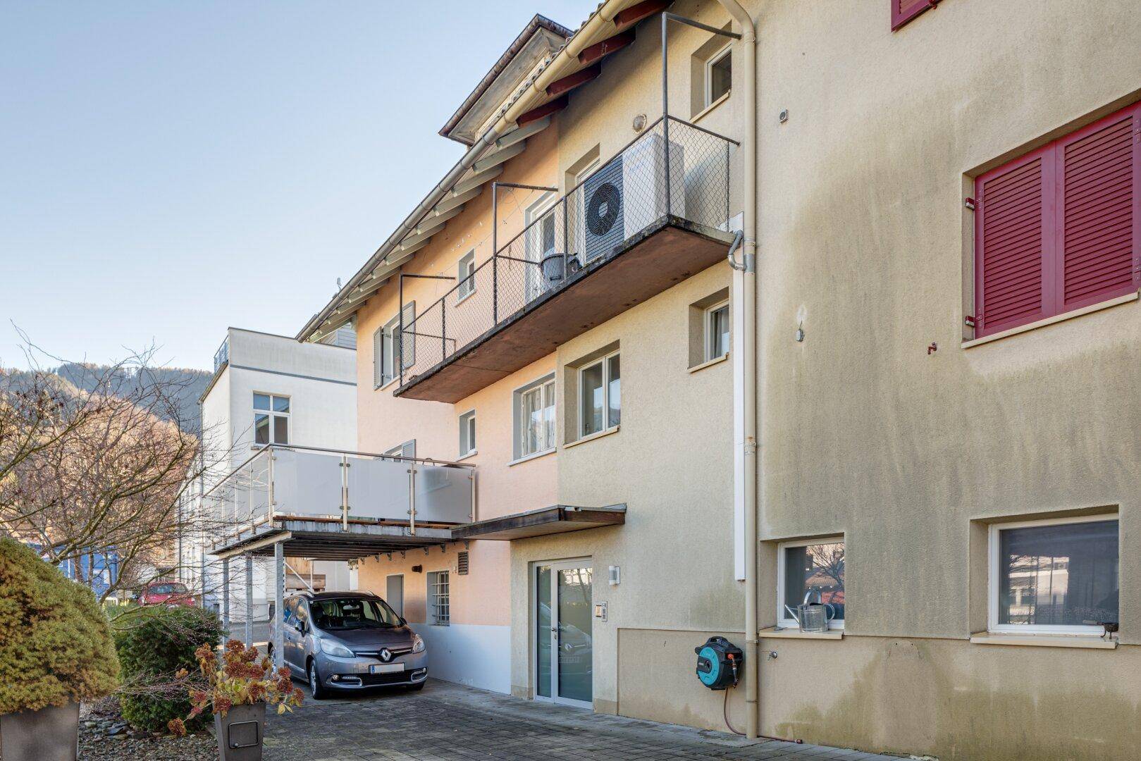 Fassade eines Wohnhauses mit mehreren Balkonen, einem geparkten Auto und roten Fensterläden.