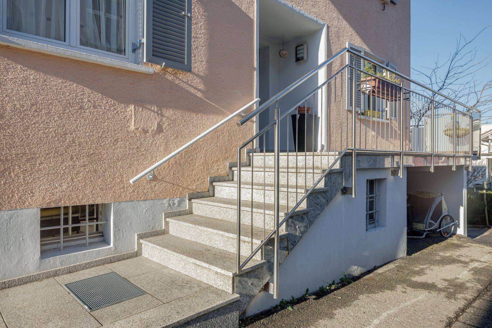 Eingangsbereich eines Gebäudes mit Steintreppe und modernem Edelstahlgeländer unter blauem Himmel.