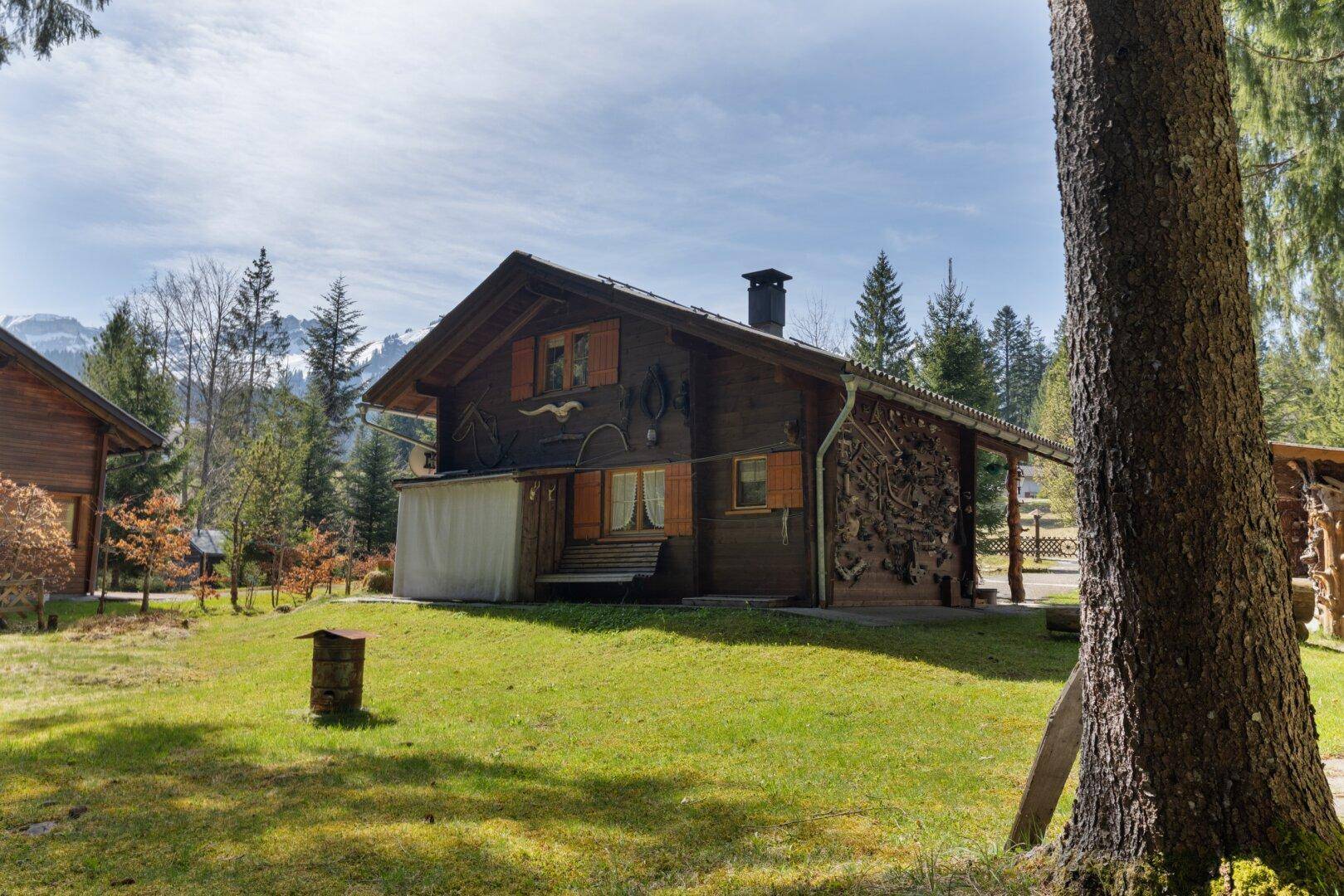 Rustikales Holzhaus mit Holzlager an der Seite und großem Gartenbereich umgeben von Bäumen.