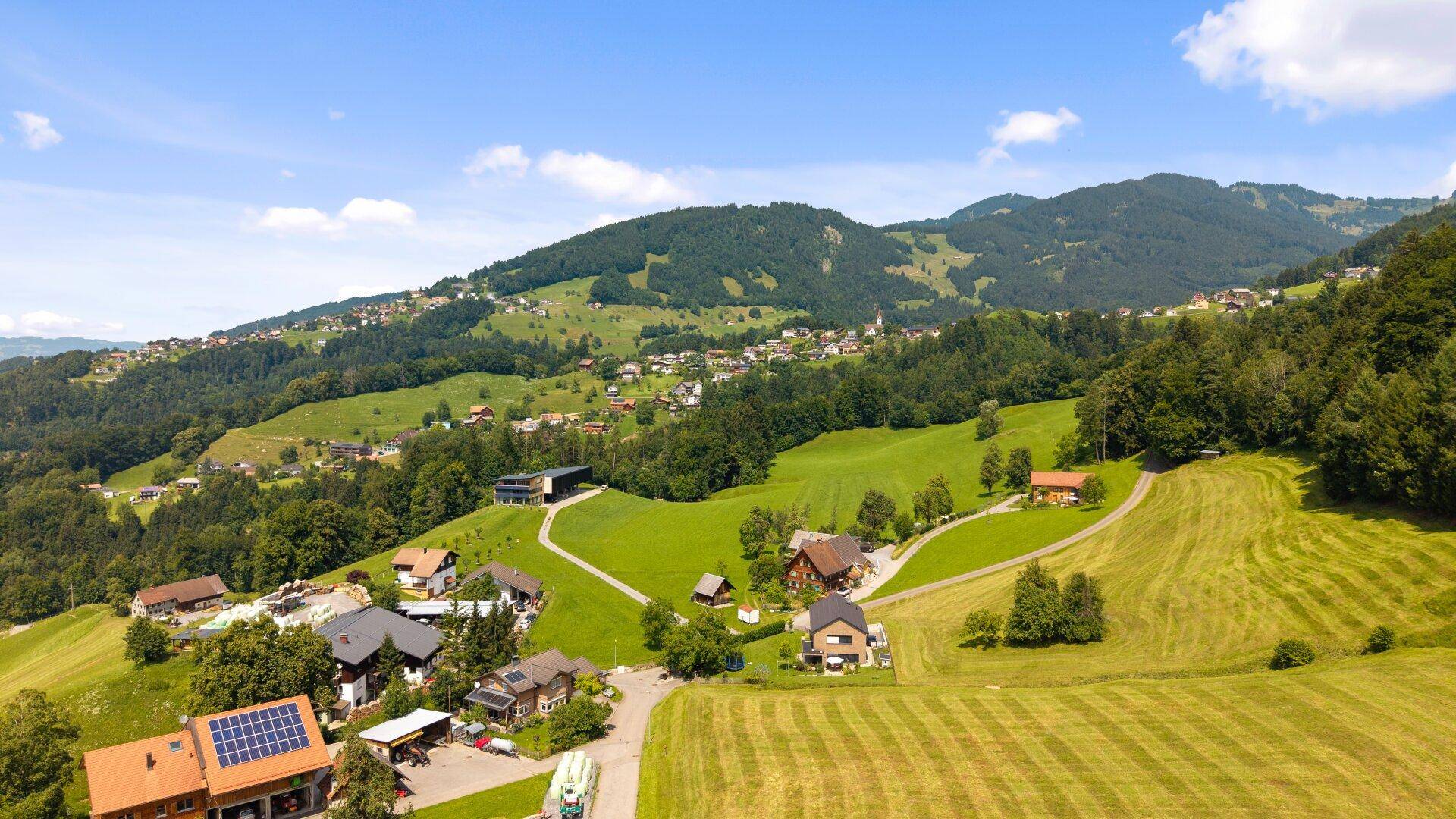 Weitläufige Luftaufnahme einer malerischen Siedlung am Hang, umgeben von Wäldern und grünen Wiesen.