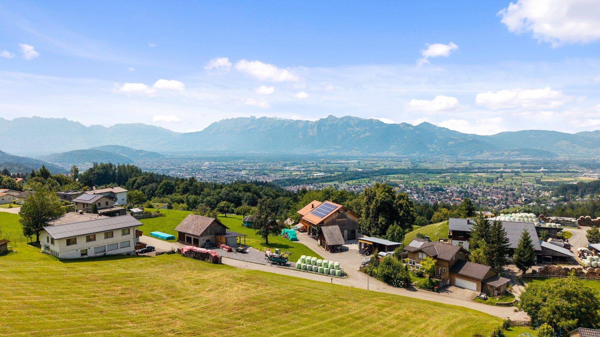 Panoramablick auf eine ländliche Siedlung mit Häusern, umgeben von grünen Hügeln und fernen Bergen.