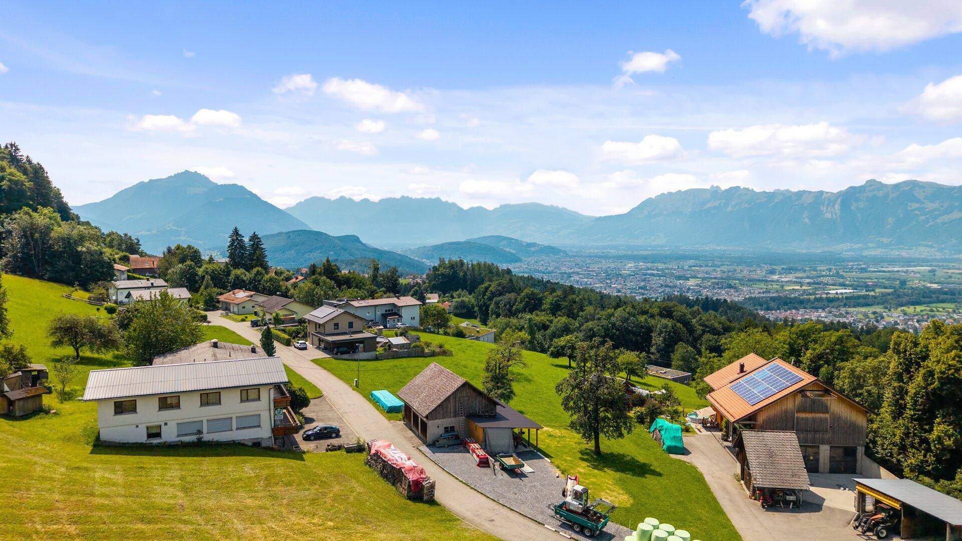 Panoramablick auf eine ländliche Siedlung mit Häusern, umgeben von grünen Hügeln und fernen Bergen.