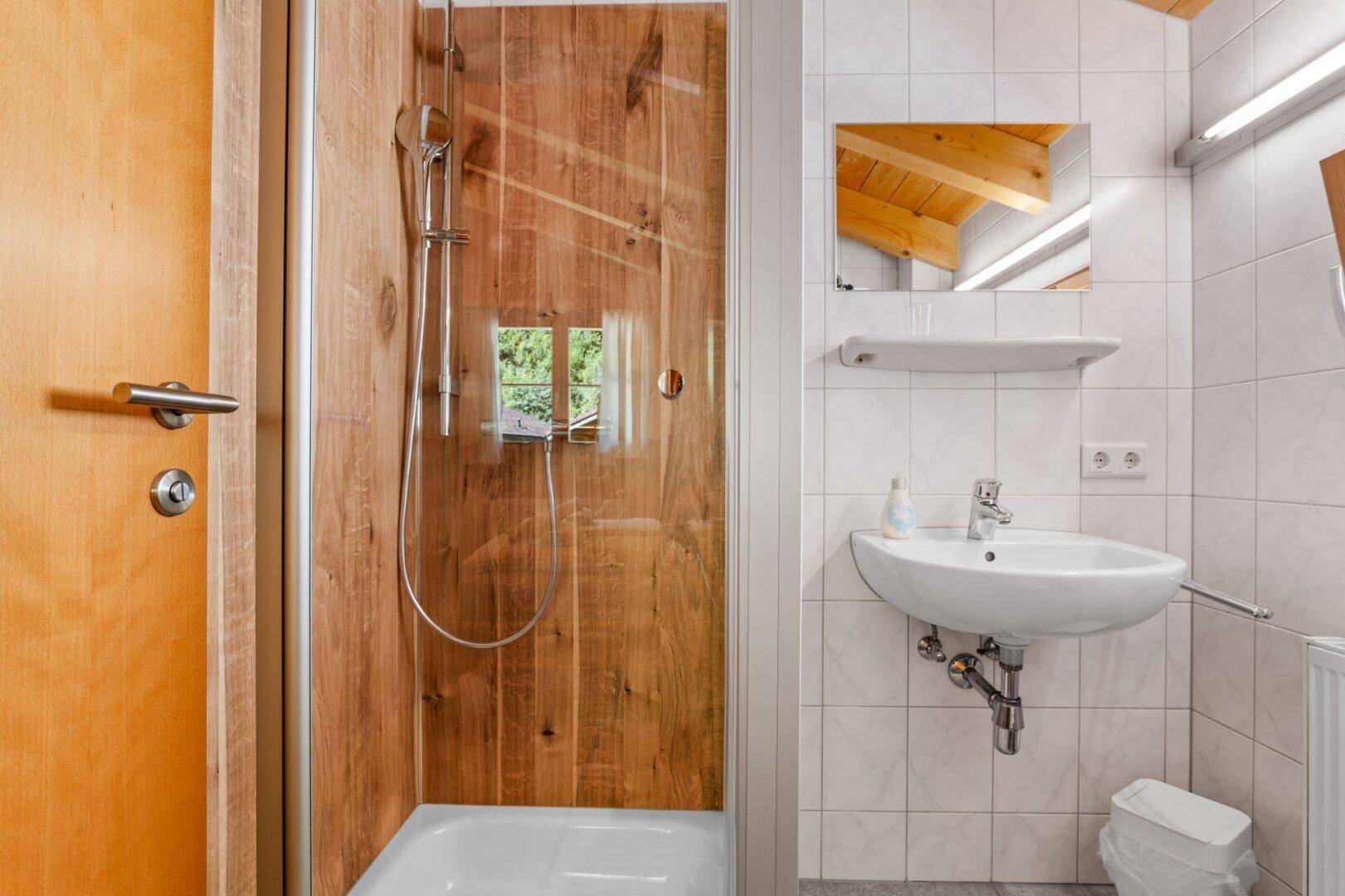 Badezimmer mit Duschkabine, Waschbecken und Spiegel, umgeben von hellen Fliesen.