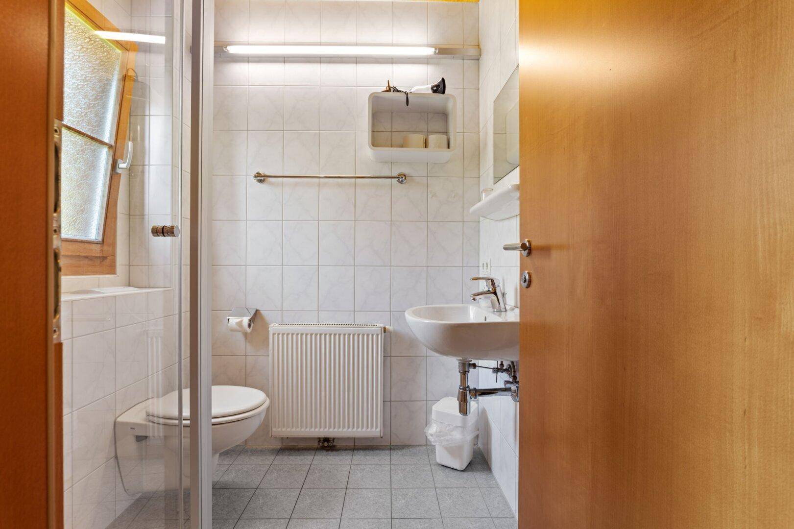 Helles Badezimmer mit Dusche, WC, Waschbecken und Heizkörper, ausgestattet mit weißen Fliesen.