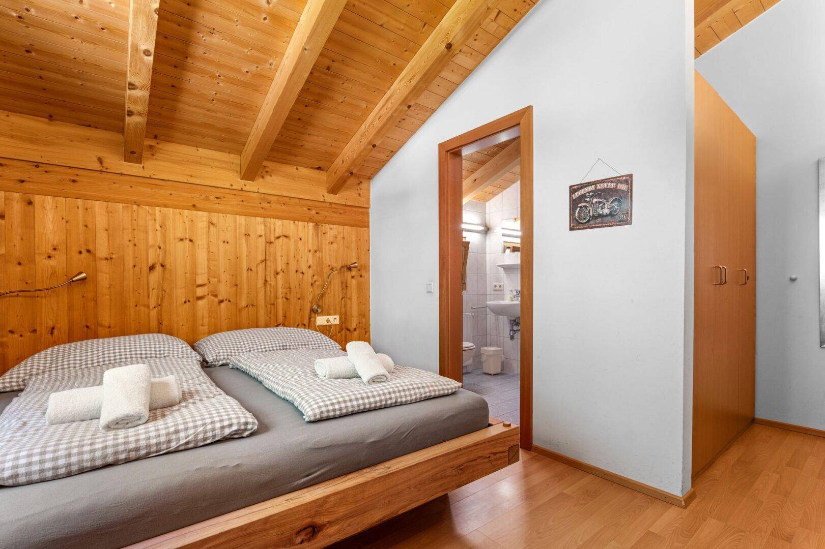 Schlafzimmer mit Holzbalkendecke, Doppelbett und Zugang zu einem angrenzenden Badezimmer.