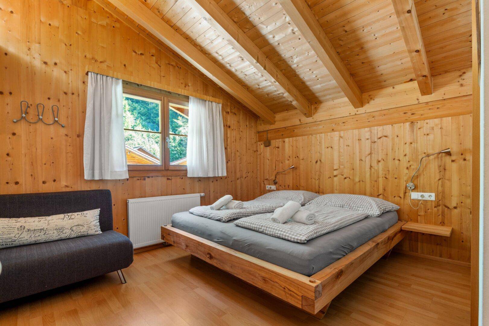Gemütliches Schlafzimmer mit Holzwänden, Holzbalkendecke und Doppelbett mit Blick ins Grüne.