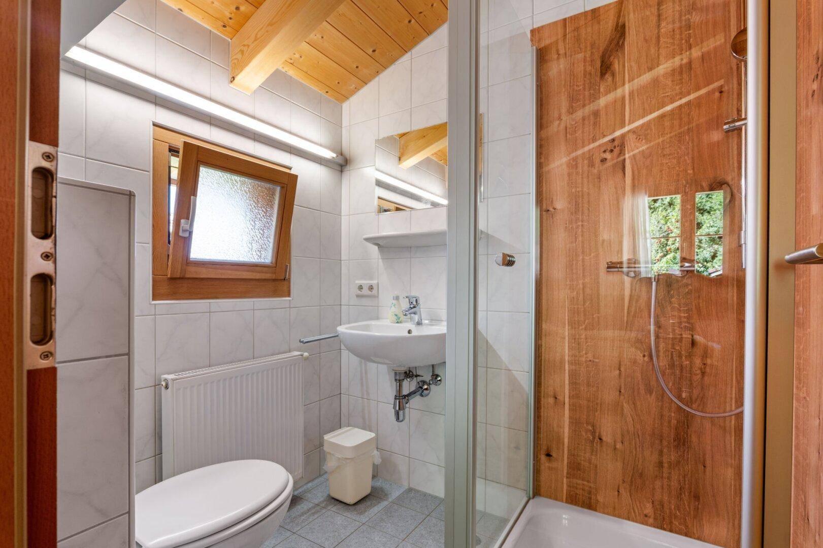 Badezimmer mit Duschkabine, WC, Waschbecken und Fenster, hell gefliest.