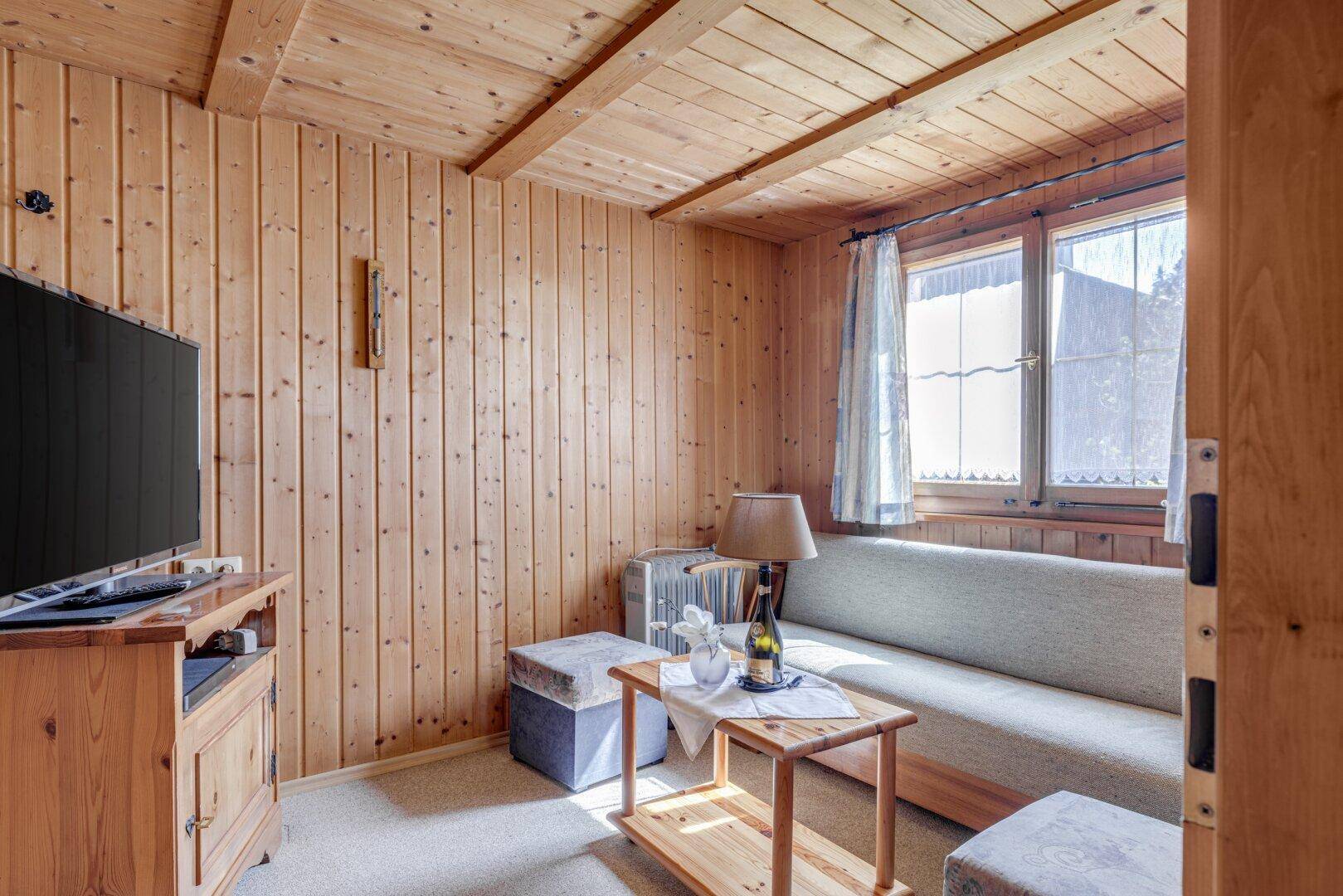 Helles Wohnzimmer mit Holztäfelung, Sofa, Fernseher und Blick ins Grüne.