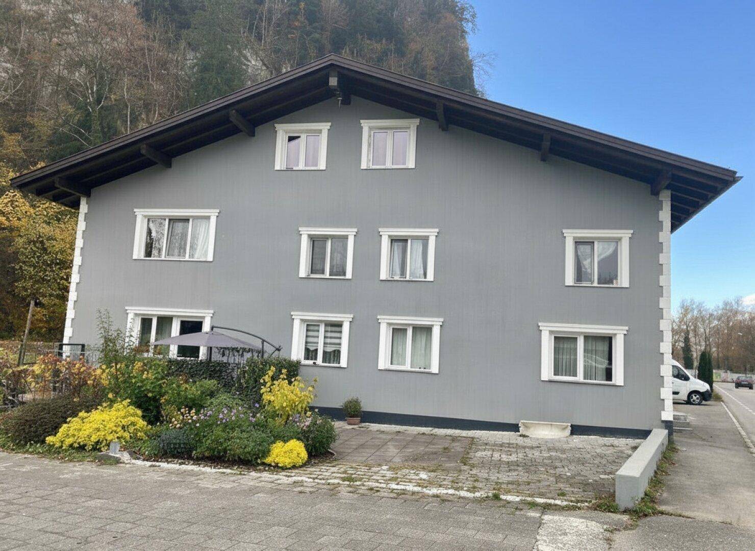 Rückansicht des grauen Gebäudes mit Gartenbereich und Blick auf bewaldete Hügel.