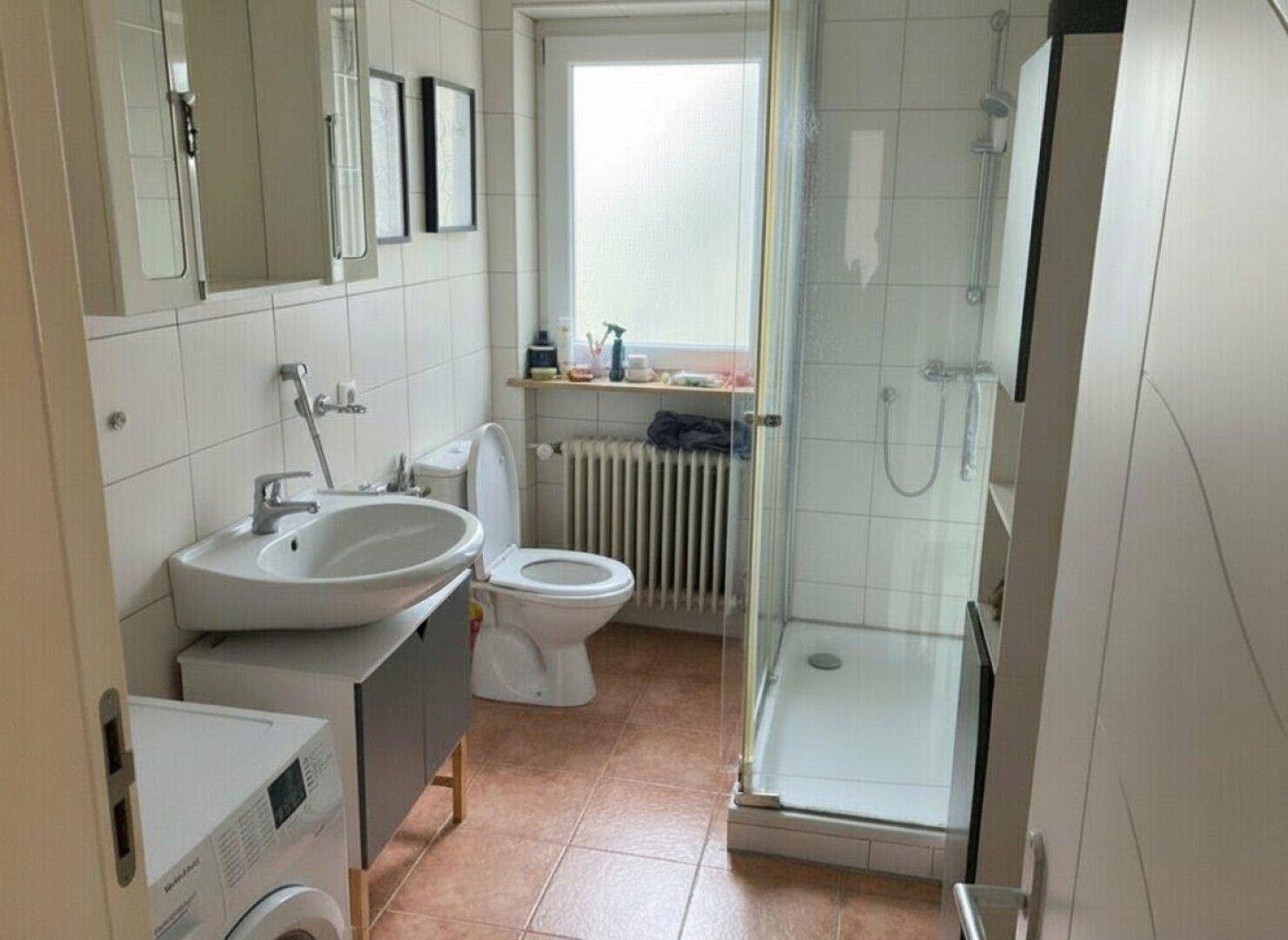 Funktionales Badezimmer mit Dusche, WC, Waschbecken und Fenster, hell gefliest.