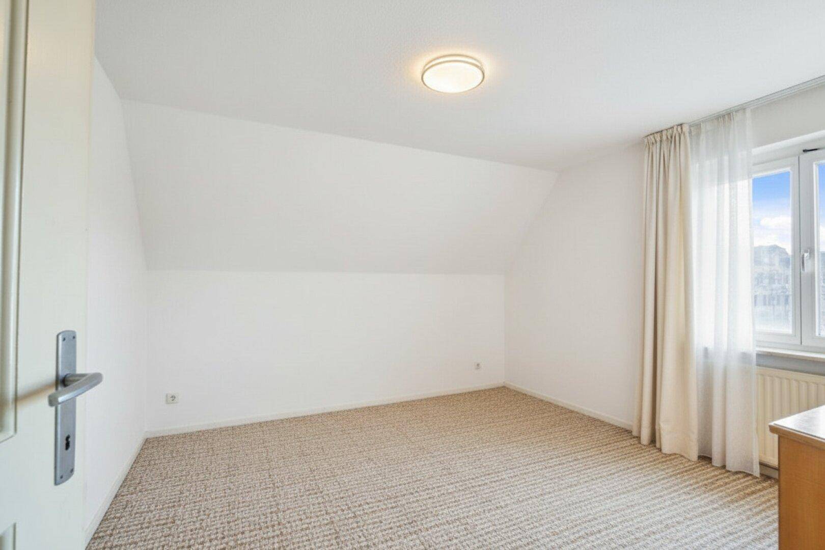 Leeres Schlafzimmer mit Teppichboden, schräger Decke und Fenster für Tageslicht.