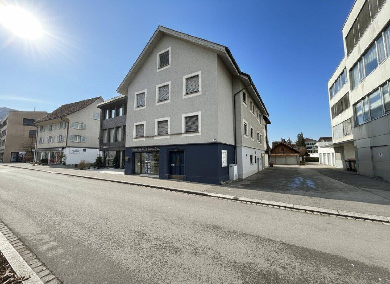 Graues Mehrfamilienhaus mit dunkelblauer Fassadenakzentuierung und Geschäften im Erdgeschoss an einer Straße.