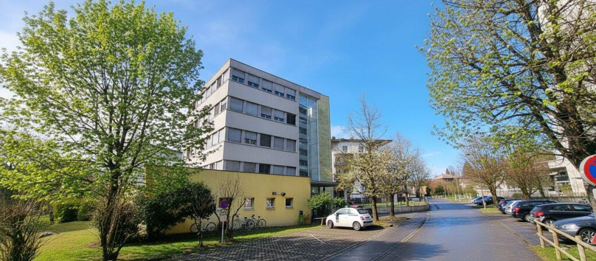 Mehrstöckiges Gebäude mit moderner Architektur, umgeben von Bäumen und einem Parkplatz unter blauem Himmel.