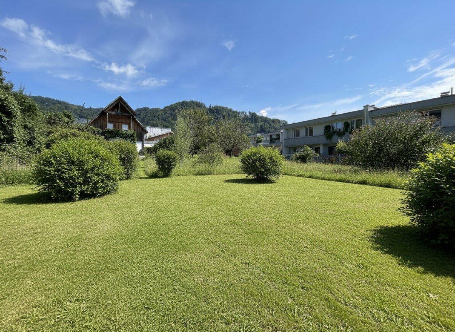 Weitläufige Rasenfläche mit Büschen und Häusern im Hintergrund, umgeben von einer Berglandschaft.