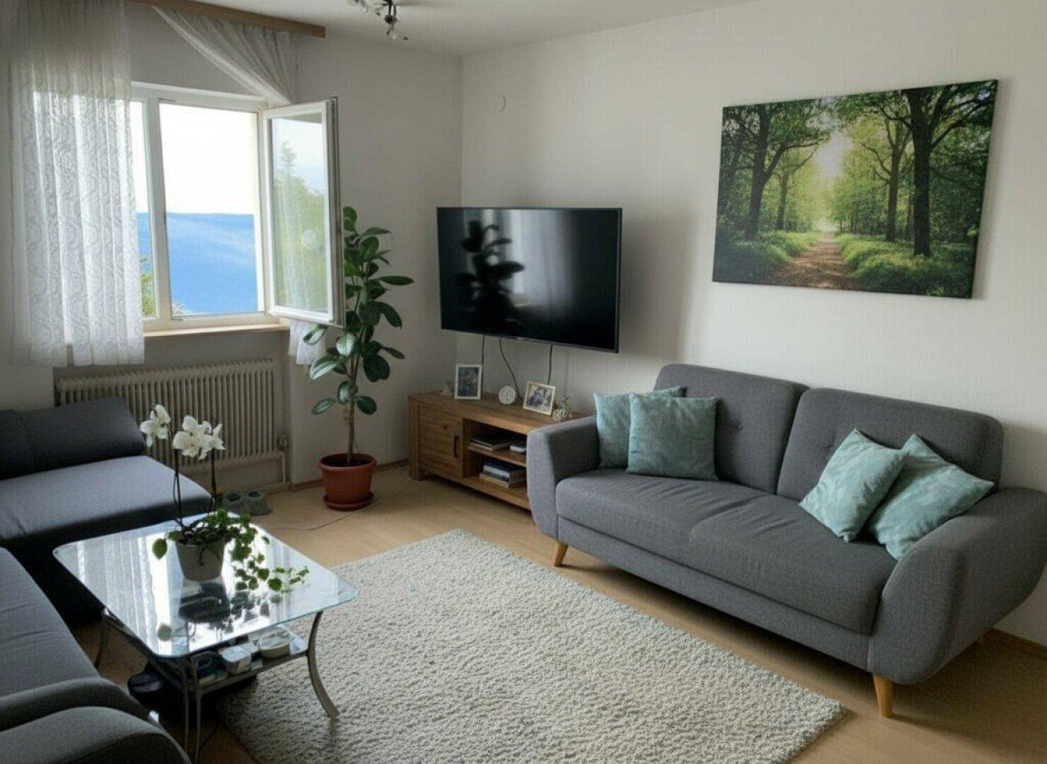 Gemütliches Wohnzimmer mit hellem Parkettboden, grauem Sofa und Blick ins Grüne.